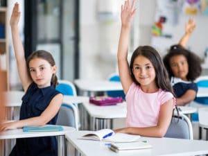 three young girls raising their hands in class